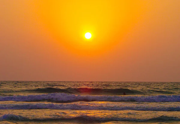 The beach at sunset is a slightly rough sea with small waves that turn into foam near the shore. The golden sun in the sky above the horizon shines in a perfect circle.