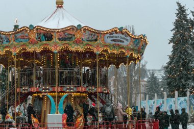Parlak ve renkli bir Noel Atlıkarınca 'sı, neşeli çelenkler ve ışıklarla parktaki açık gökyüzünün altında duruyor. Atlıkarıncanın etrafında neşeli bir sürü insan var..