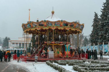 Parkın ortasında, açık gökyüzünün altında, neşeli çelenkler ve ışıklarla dolu bir Noel Atlıkarınca 'sı. Atlıkarıncanın etrafında neşeli bir sürü insan var..