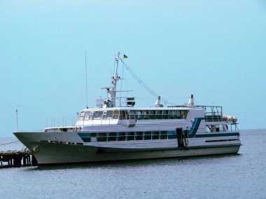 Gemi küçük bir körfezin yanında demirli. Pürüzsüz mavi deniz suyu ve açık, güneşli gökyüzü.
