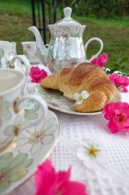 Çiçek desenli çaydanlık, çay fincanları, Çin 'de altın kruvasan ve beyaz dantelli kumaş üzerinde taze pembe çiçekler olan güzel bir açık hava çay masası.