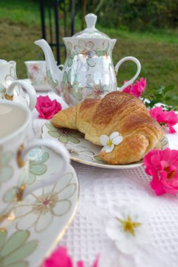 Çiçek desenli çaydanlık, çay fincanları, Çin 'de altın kruvasan ve beyaz dantelli kumaş üzerinde taze pembe çiçekler olan güzel bir açık hava çay masası.