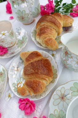 Çiçek desenli çaydanlık, çay fincanları, Çin 'de altın kruvasan ve beyaz dantelli kumaş üzerinde taze pembe çiçekler olan güzel bir açık hava çay masası.