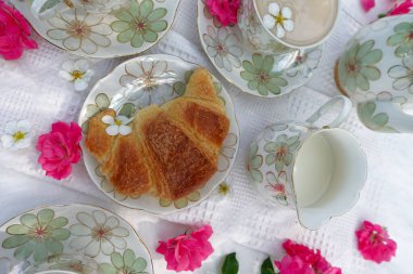 Çiçek desenli çaydanlık, çay fincanları, Çin 'de altın kruvasan ve beyaz dantelli kumaş üzerinde taze pembe çiçekler olan güzel bir açık hava çay masası.