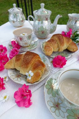 Çiçek desenli çaydanlık, çay fincanları, Çin 'de altın kruvasan ve beyaz dantelli kumaş üzerinde taze pembe çiçekler olan güzel bir açık hava çay masası.