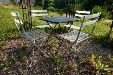 Beyaz metal sandalyeler ve pergola altında bir masa ile huzurlu bir açık hava oturma alanı, uzak tepeleri ile yemyeşil bir manzara.