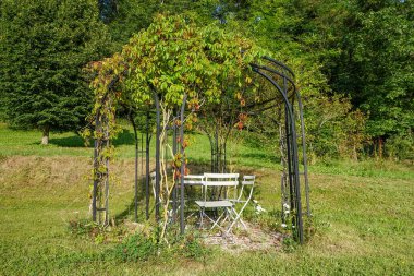 Beyaz metal sandalyeler ve pergola altında bir masa ile huzurlu bir açık hava oturma alanı, uzak tepeleri ile yemyeşil bir manzara.