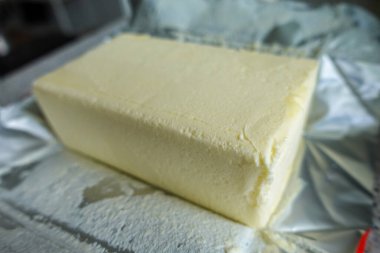 Açık folyo ambalajının üzerinde duran dikdörtgen bir tereyağı bloğu kremsi ve pürüzsüz bir dokuyu gösteriyor..