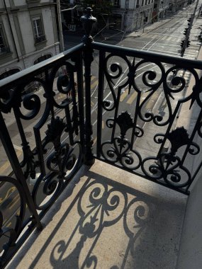 Karmaşık işlenmiş demir bir balkon korkuluğu taş zeminde zarif gölgeler bırakıyor. Tramvay rayları ve yaya geçitleri olan sakin bir şehir caddesine bakıyor.. 