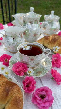 Zarif çiçek porselenleri, taze kruvasanlar ve canlı pembe güller içeren güzel bir açık hava çay partisi büyüleyici ve davetkar bir atmosfer yaratıyor..