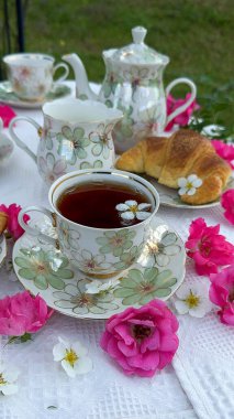 Zarif çiçek porselenleri, taze kruvasanlar ve canlı pembe güller içeren güzel bir açık hava çay partisi büyüleyici ve davetkar bir atmosfer yaratıyor..