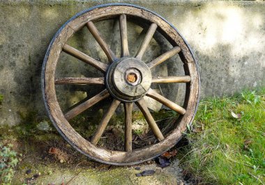 Yaşlı, yosun kaplı beton bir duvara yaslanmış kırsal ahşap bir vagon tekerleği. Tekerlek tahtası görünür şekilde aşınmış ve çatlamış, merkezi metal merkezinde pas var.