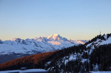 Batan güneşin altın parıltısıyla yıkanan karla kaplı dağ tepelerinin çarpıcı manzarası. Aydınlatılmış tepeler ve kararan gökyüzü arasındaki zıtlık nefes kesici bir alp manzarası yaratır..