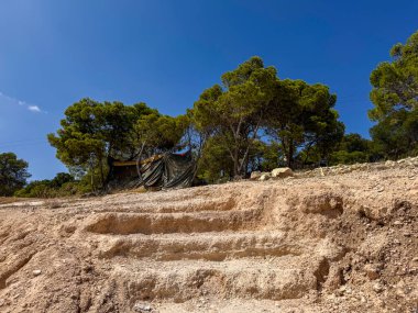 Muşambadan yapılmış engebeli bir orman sığınağı, canlı mavi bir gökyüzünün altında, çam ağaçlarıyla çevrili engebeli taş basamakların üzerinde duruyor..