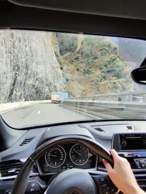 Sürücüler, dramatik bulutların ve mavi gökyüzünün altında, koruyucu bariyerlerle kıvrımlı bir otoyolda ilerleyen bir arabanın içinden görüyorlar..