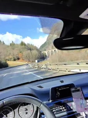 Mavi gökyüzü, uçurumlar ve orman manzarasıyla çerçevelenmiş uzun bir taş viyadük altında kıvrılan bir dağ yolu manzarası..