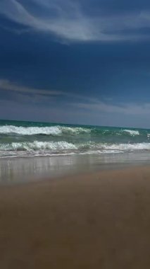 Berrak bir dalga, parlak güneş ışığı altında, temiz, altın gibi kumlu bir plaja doğru yavaşça yuvarlanıyor. Sakin ve yaz atmosferini anımsatıyor..