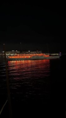 Güzel aydınlatılmış bir tekne, gece karanlığında altın ve kırmızı ışıklarını yansıtır. Işıldayan yansımalar etrafındaki karanlıkla çarpıcı bir kontrast oluşturuyor..