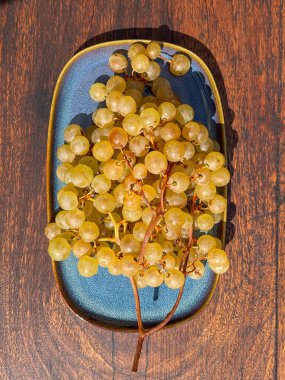 Tahta bir yüzey üzerinde mavi bir tabağa yerleştirilmiş olgun beyaz üzümler. Tazeliği, hasatı ve doğal tatlılığı simgeliyor..