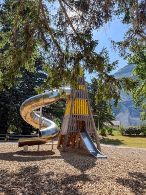 Güneşli bir günde ağaçlar ve dağlarla çevrili yüksek sarmal bir oyun alanı. Çocukluk eğlencesini, açık hava eğlencesini ve macerayı simgeliyor..