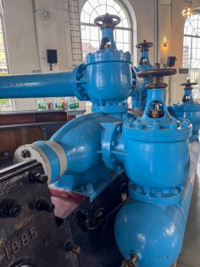Kapakçıkları ve boruları olan büyük mavi endüstriyel makineler kemerli pencereleri ve detaylı tavan mimarisi olan tarihi bir binanın içinde sergileniyor..