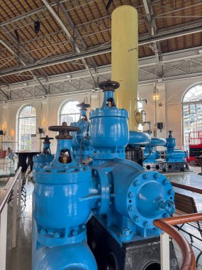 Kapakçıkları ve boruları olan büyük mavi endüstriyel makineler kemerli pencereleri ve detaylı tavan mimarisi olan tarihi bir binanın içinde sergileniyor..