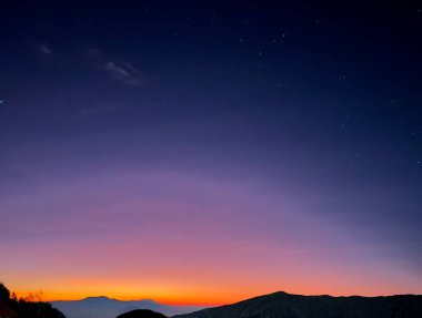 Ufukta parlak, turuncu ve pembe bir günbatımı olan yıldızlı gece gökyüzünün manzarası. Siluetli dağlar görüntünün alt köşesinde dramatik ve barışçıl bir manzara yaratıyor..