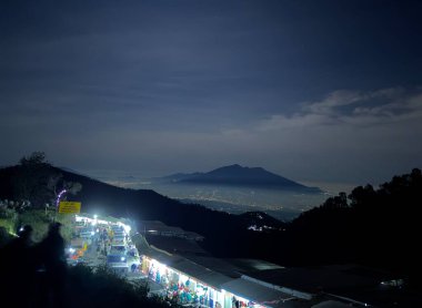 A night market stretches along a hillside, illuminated by string lights. In the distance, a mountain silhouette rises against a starry sky with clouds. clipart