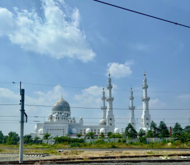 Birden fazla kubbe ve minaresi olan beyaz bir cami mavi gökyüzü ve tüylü bulutların önünde duruyor. Cami yeşilliklerle çevrili ve ön planda demiryolu rayları var..