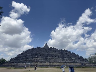 Endonezya 'daki Borobudur Tapınağı manzarası. Devasa taş yapı karmaşık oymalarla kaplı ve üzerinde sayısız taş var. Gökyüzü parlak bir mavi ve kabarık beyaz bulutlar.