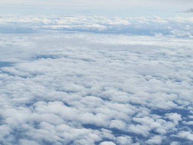 Bir uçak penceresinden panoramik bir manzara gökyüzünde uzanan tüylü beyaz kümülüs bulutlarının engin genişliğini gösteriyor. Bulutlar sıra sıra dizilmiş gibi görünüyor, büyüleyici bir desen oluşturuyor..