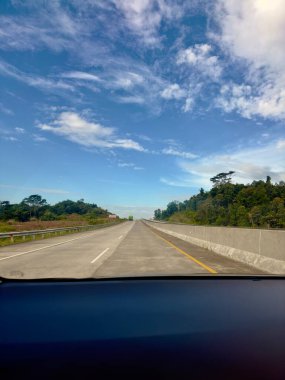 A view from a car driving on a highway. The road stretches into the distance, bordered by a concrete barrier and trees on either side. The sky is a bright blue with white fluffy clouds. clipart