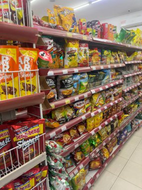 Market koridorunda cipsler, kurabiyeler ve krakerler dahil çeşitli abur cuburlar var. Raflar düzenli ve iyi aydınlatılmış, çeşitli marka ve tatlar sergileniyor..
