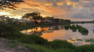 Gün batımının canlı renkleri sakin bir göle yansıyor. Ağaçlar ve yapılar kıyı boyunca uzanır ve sakin bir manzara yaratır. Gökyüzü dramatik bulutlar sergiliyor.