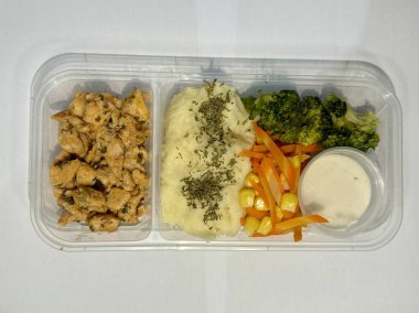 Plastik tepside hazırlanmış bir yemek ızgara tavuk, patates püresi, buğulanmış brokoli, havuç, mısır ve kremalı sos içerir. Dengeli ve uygun bir yemek seçeneği..