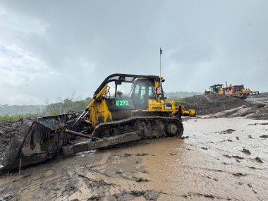 Güçlü sarı bir buldozer, bulutlu bir gökyüzünün altında çamurlu bir inşaat alanında ağır makineler ve yer değiştirme operasyonlarını zorlu bir hava koşulunda gösteriyor..