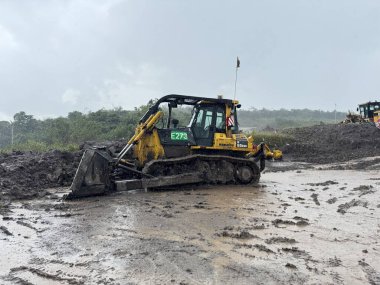 Ağır makineler çamurlu bir inşaat alanını ele geçiriyor. Sarı bir buldozer gri bir gökyüzünün altında ıslak toprakta manevra yapıyor. Kötü koşullar altında güçlü ve dirençli..