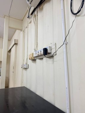Endüstriyel elektrik kablolarının, prizlerin ve elektrik bantlarının yakın görüntüsü koyu bir tezgahın bitişiğindeki bej bir duvara monte edilmiş. İnşaat altyapısını resmediyor.
