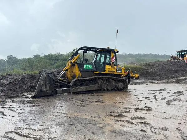 Ağır makineler çamurlu bir inşaat alanını ele geçiriyor. Sarı bir buldozer gri bir gökyüzünün altında ıslak toprakta manevra yapıyor. Kötü koşullar altında güçlü ve dirençli..