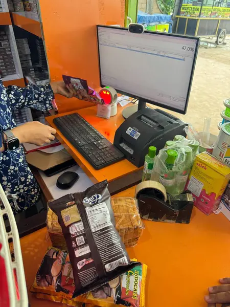 Bir kasiyer, canlı bir portakal kasasında bilgisayar kullanarak bir market alışverişi yapar. Çeşitli atıştırmalıklar ve içecekler görünür.