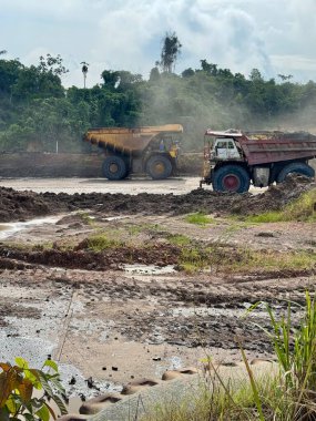 Güçlü maden damperli kamyonlar, verimli yeşil bitki örtüsünün yakınındaki engebeli bir arazide doğal ortamdaki endüstriyel ağır makineleri gösteren bulutlu bir gökyüzünün altında maden cevheri taşıyorlar..