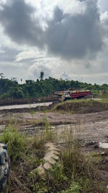 Bulutlu gökyüzünün altındaki inşaat sahasında verimli yeşil bitki örtüsü yakınlarında toprak kazan ağır makineler bulunuyor..