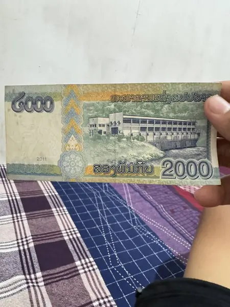 Eskimiş bir 2000 Lao Kip banknotu sergilenmektedir. Tasarıda bir bina ve su görüntüsü yer alıyor. Kumaş arka planına karşı bir el tarafından tutuluyor..