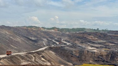 Geniş açı parçalı bulutlu bir gökyüzü altında devasa bir açık maden operasyonu yakalar. Ağır makineler, engin ve çorak arazide kazılmış materyalleri taşıyarak görülebilir..