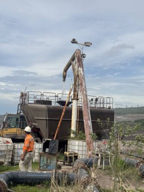 Bulutlu gökyüzü altında büyük makineler ve boru hatları yakınında sanayi işçisi