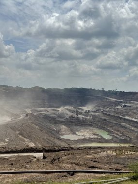 Dramatik bulutlu bir gökyüzü altında engin endüstriyel madencilik sahası. Toprak çıkarma ve ağır makineler çalışıyor.