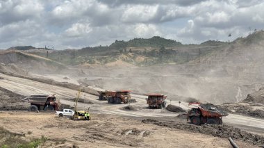 Massive mining trucks haul coal across dusty landscape under dramatic cloudy sky, industrial extraction process in action