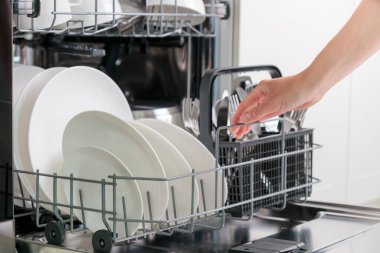 Kadın eli bulaşık makinesini sofra takımıyla dolduruyor. Mutfak aletleri, yaşam tarzı manzarası. Kadın bulaşık makinesinden çatal bıçak sepetini alıyor. Mutfaktaki modern teknoloji kavramı