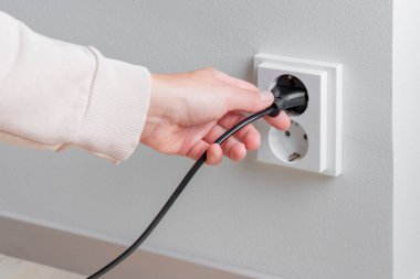 Beyaz bir duvardaki elektrik prizinden elektrik kablosunu söken bir kadın eli. Dikkatli kaynak kullanımı, artan elektrik maliyeti ve günlük hayatta dikkatli tüketim, güvenlik ve sorumlu enerji yönetimi ihtiyacı