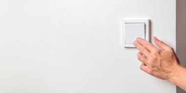 Beyaz bir duvardaki ışık düğmesini kapatan bir kadının eli. Elektrik tasarrufu ve dikkatli kaynak kullanımı, artan elektrik maliyetlerinin etkisi ve dikkatli enerji yönetimi ihtiyacı